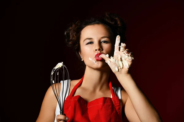 Young woman chef cook hold wire whisk eggs beater for cup cakes with cream and lick finger on dark red