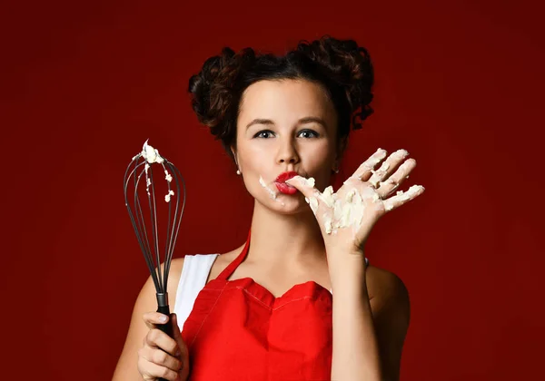 Young woman chef cook hold wire whisk eggs beater for cup cakes with cream and lick finger on dark red