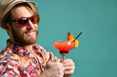 Genç adam şapka Haritayı başparmak kırmızı margarita kokteyl içki içecek suyu mutlu kameraya mavi üzerinde seyir