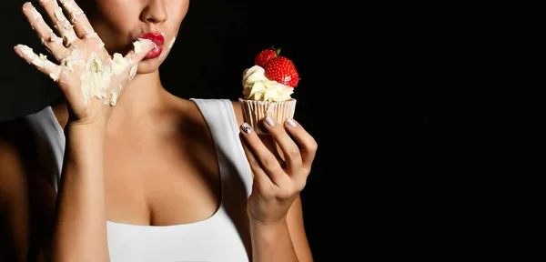Молода жінка кухаря тримає солодкий чашковий торт з вершками полуниці лизати палець на чорному — стокове фото