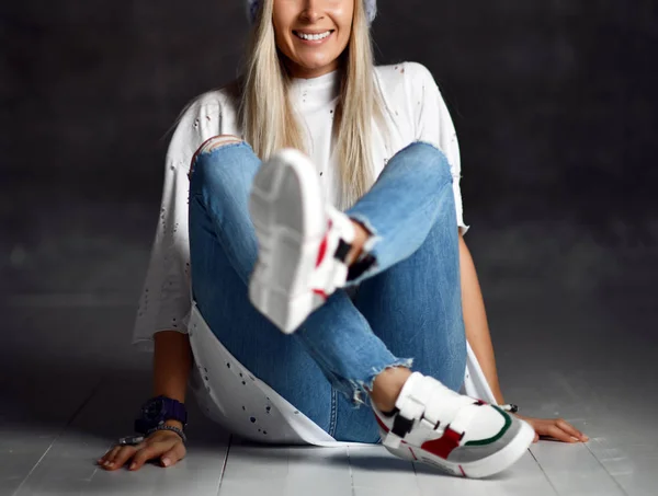 Blonde vrouw in lichte blauwe gebreide muts jeans en witte t-shirt gelukkig lachend zittend op houten vloer — Stockfoto