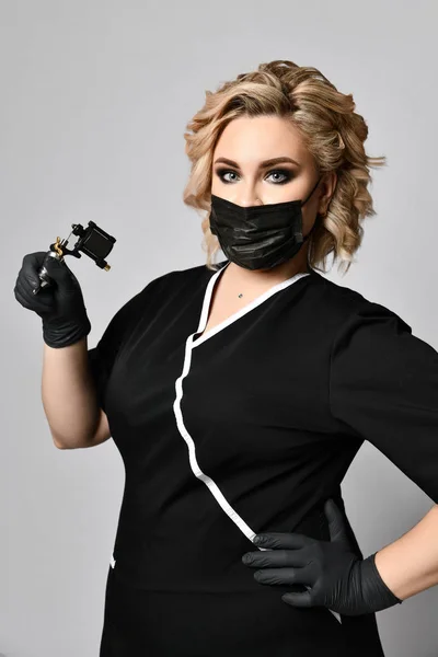 Жінка-косметолог тримає татуювальний пістолет у чорній масці на сірому — стокове фото