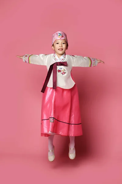 Bambina coreana in abito tradizionale di seta per festeggiare il nuovo anno ballando saltando sul rosa con le mani distese — Foto Stock