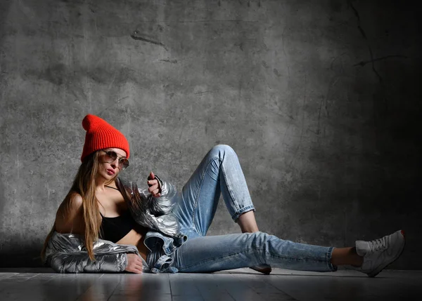 Young sport woman after workout exercise sitting point finger in silver sexy thin down puffer jacket blue jeans red hat