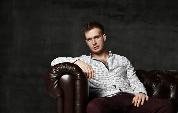 Young business man sitting in leather Chester armchair sofa in casual shirt thinking on dark background