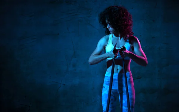 Female fitness instructor working out with a rubber resistance band in blue and pink neon light in gym