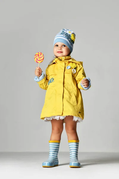 Yürümeye başlayan çocuk kız çocuk büyük tatlı lolipop şeker sarı ceket ve lastik çizme ile genç güzel — Stok fotoğraf