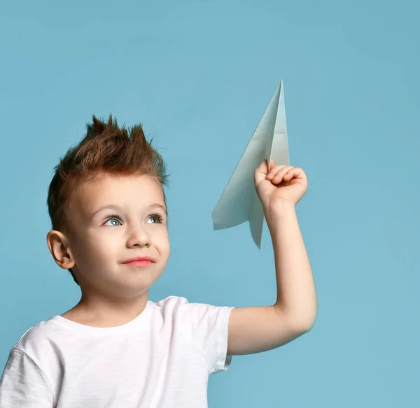 Jovem menino menino em branco t-shirt segurar o origami avião de papel sorrindo olhando para cima em azul claro — Fotografia de Stock