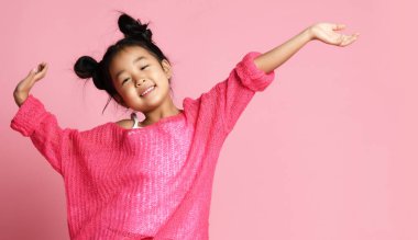 Asian kid girl in pink sweater, white pants and funny buns stands with hands up and smiles on pink background. close up