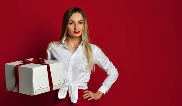 Młoda blond kobieta trzymać białe pole prezent obecny uśmiechający się spróbować go otworzyć na urodzinowe party na czerwono — Zdjęcie stockowe