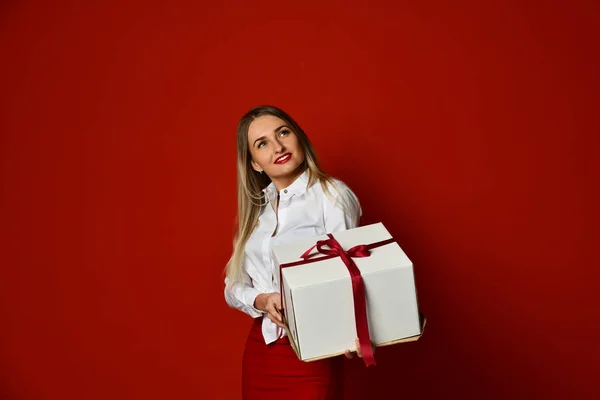 Giovane donna bionda tenere scatola bianca regalo sorridente cercare di aprirlo sulla festa di compleanno celebrazione sul rosso — Foto Stock