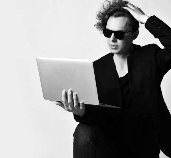 Giovane uomo moderno in occhiali da sole guarda qualcosa sul computer portatile che tiene sul ginocchio e graffia la testa pensieroso . — Foto Stock