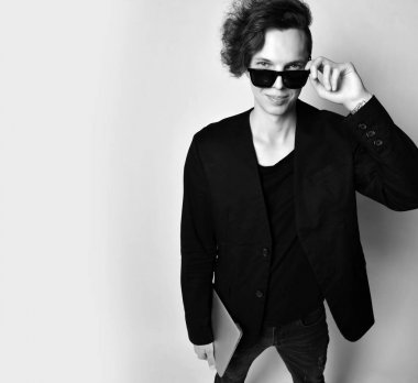 Siyah tişörtlü, ceketli ve kotlu genç bir adam dizüstü bilgisayarlı şık güneş gözlüğü ve gülüşünün üstünde bize bakıyor. Siyah ve beyaz.