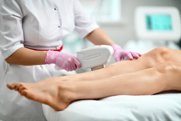 Primer plano. Cosmetólogo en bata blanca y guantes realiza un procedimiento en pierna paciente con equipo médico láser en salón de belleza equipado —  Fotos de Stock