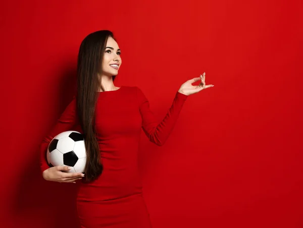 Mujer morena bastante sonriente en vestido rojo apretado con pelota de fútbol en la mano mira hacia arriba soñadoramente y cuenta sus dedos — Foto de Stock