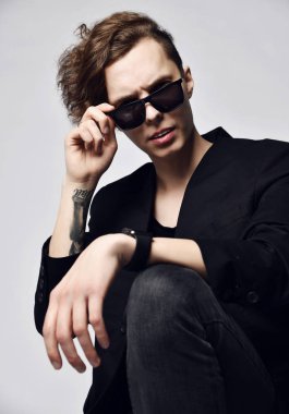 Yaratıcı saç kesimi ve elinde dövmesi olan genç şık bir adam yüksek bir sandalyede oturuyor ve güneş gözlüklerini izliyor.