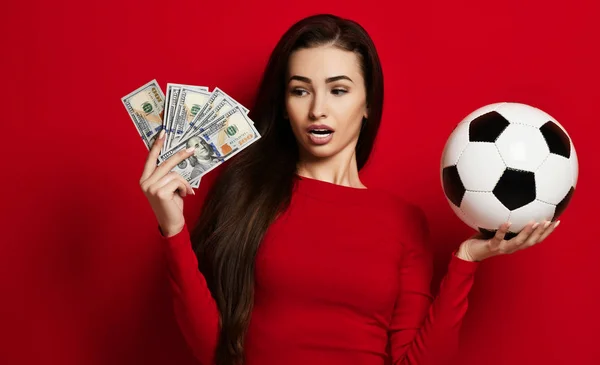 Молода брюнетка в жорсткій червоній сукні тримає футбольний м'яч в одній руці і здивована пучками доларових банкнот в іншій — стокове фото