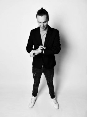 Modern saç kesimi gri balıkçı yaka, siyah ceket, kot pantolon ve spor ayakkabı bir topuz ile şık adam onun saat bakar. Siyah beyaz.