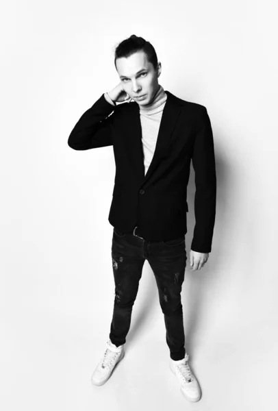 Homem elegante jovem com um pão moderno na cabeça em jaqueta preta, jeans e tênis brancos ajusta colarinho de sua gola de gola alta cinza — Fotografia de Stock