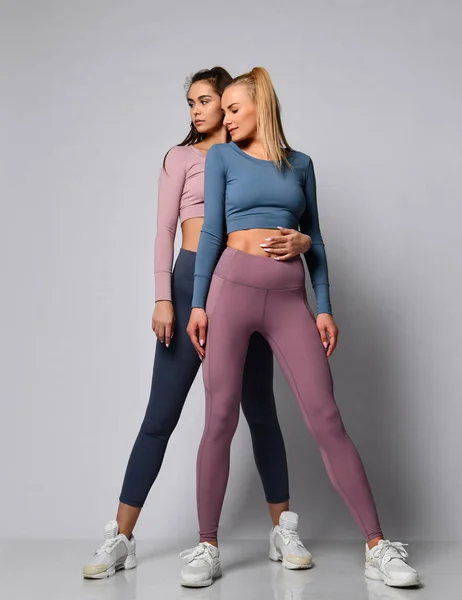 Two sporty blonde and brunette girls in athletic body cloth sport wear cloth stand together after workout on gray — Stock Photo, Image