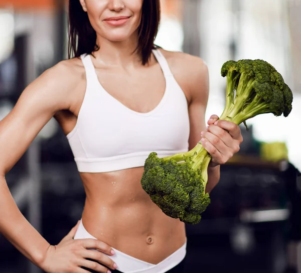 Gros plan. Régime alimentaire et concept de perte de poids. Moniteur de fitness femme athlétique en vêtements de sport avec un gros brocoli comme poids dans la salle de gym — Photo