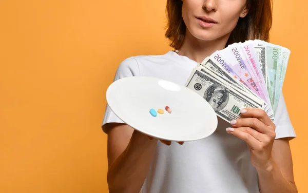 Жінка-дієтолог тримає тарілку з різними кольоровими таблетками таблетки дієтичні добавки рецептурні ліки та долари гроші валюта на жовтому — стокове фото