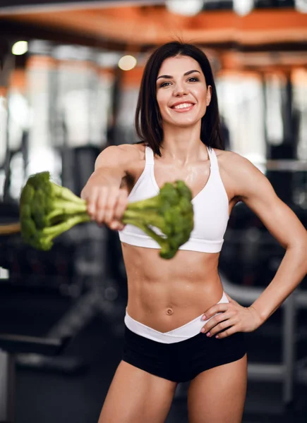 Koncepce výživy a úbytku váhy. Veselý, usměvavý instruktor fitness dívky nám podal velkou brokolici jako závaží. — Stock fotografie