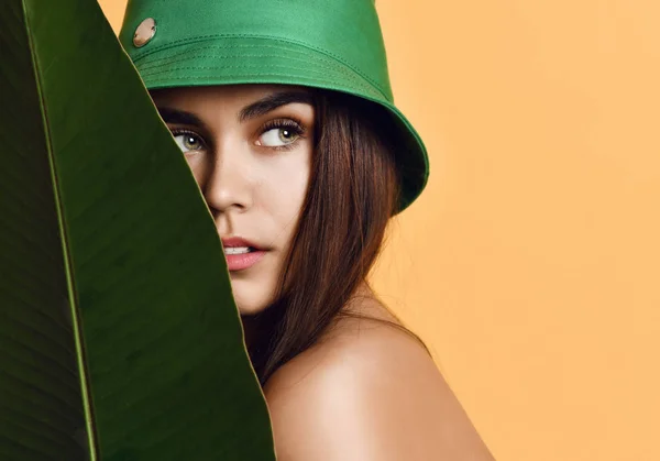 Vacker ung mode flicka med perfekt hud i grön hatt hålla tropiska bananblad i händer och omslag — Stockfoto