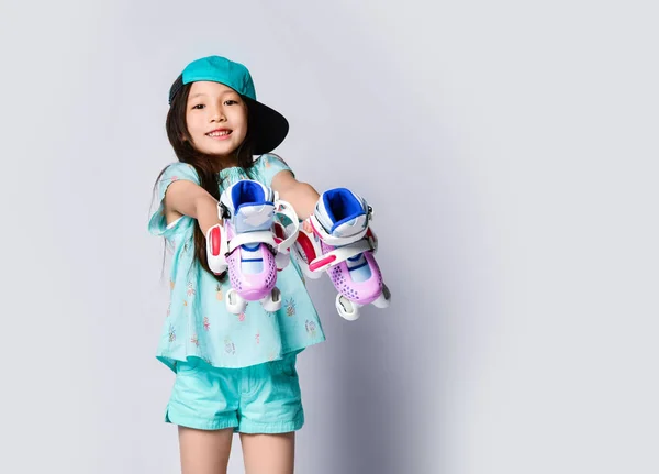 Маленька азіатська дівчинка дитина сидить з роликовими ковзанами у світло-блакитній футболці та шапці щаслива усміхнена на білому — стокове фото