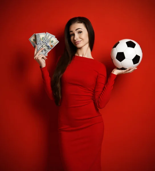 Jonge slyly glimlach brunette vrouw in strakke rode jurk houdt voetbal en bundels van dollars nota — Stockfoto