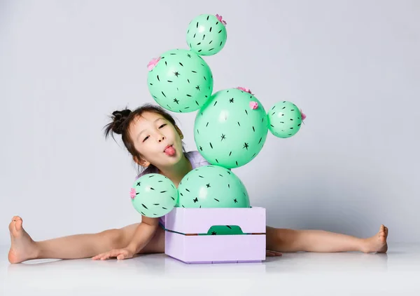 Смішна азіатська дівчина сидить за коробкою з кактусовою кулею, виглядає і застряг її язиком — стокове фото