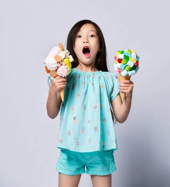 Asian baby girl kid holds two sweet big ice-cream in waffles cones with tasty marshmallow jelly beans can not believe her luck clipart