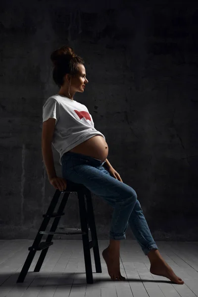 Hermosa joven embarazada feliz madre mujer en camiseta blanca posando en la oscuridad —  Fotos de Stock