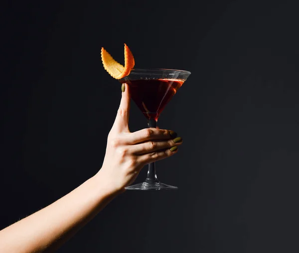 Young woman hand with a delicious red cocktail drink with orange in martini glass on dark — Stock Photo, Image