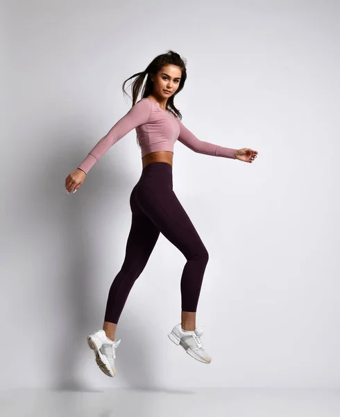 Young athletic woman brunette girl in a good shape with hair in a ponytail in trendy  sportswear gym uniform jumps dances