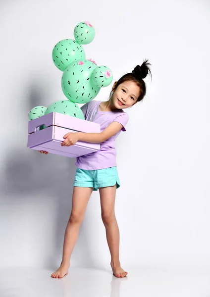 Smiling asian girl kid carries and peeks out from behind a blue wooden flower box with a cactus balloon with pink flowers in it — Stock Photo, Image