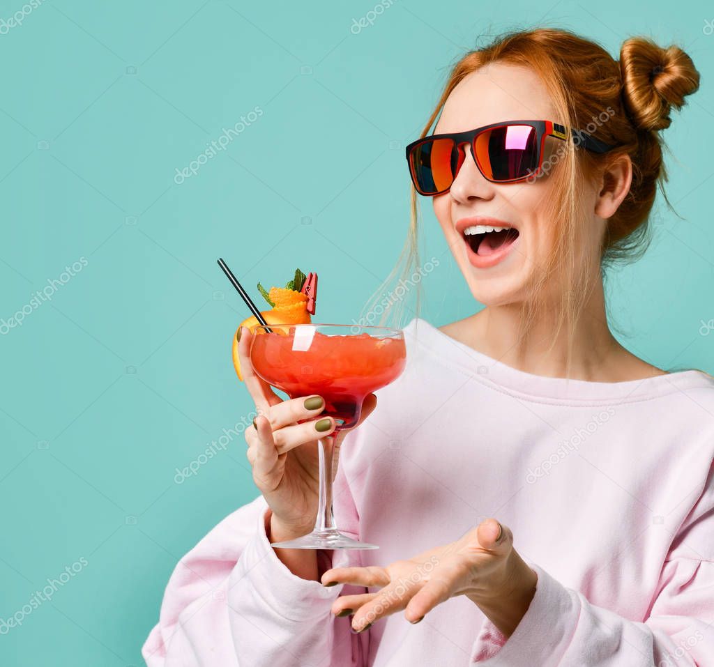 Joyful blonde girl in modern red sunglasses with tropic cocktail strawberry margarita has fun chatting with friends gesturing