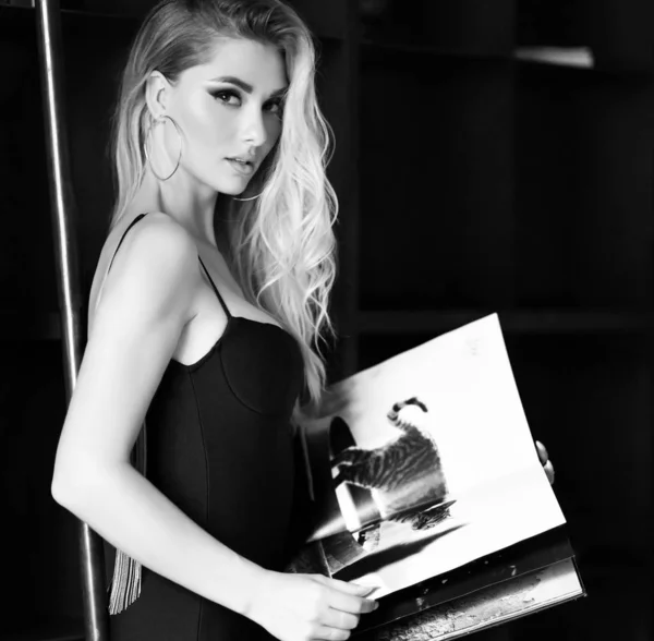Young pretty woman in sexy little black dress reading magazine or book in modern room — Stock Photo, Image