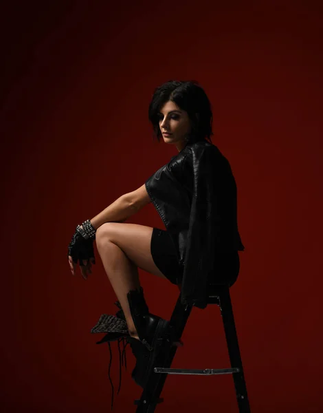 Zamyšlená žena v černých těsných šatech, kožené bundě a velkých brutálních botách, které sedí na šátku a dívá se dolů na červenou — Stock fotografie