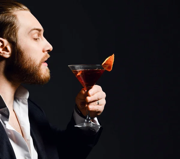 Молодий бізнесмен бородатий клубник у білій сорочці і куртці насолоджується запахом червоного коктейльного напою з апельсином у мартіні склянці — стокове фото