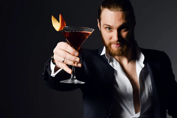 Young modern man bearded barman in fashion suit offers to try a delicious red cocktail drink with orange in martini glass