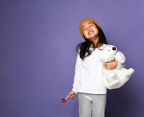 Счастливая улыбающаяся смеющаяся азиатская девочка держит игрушку белого медведя и конфетку с леденцом на синей мяте — стоковое фото
