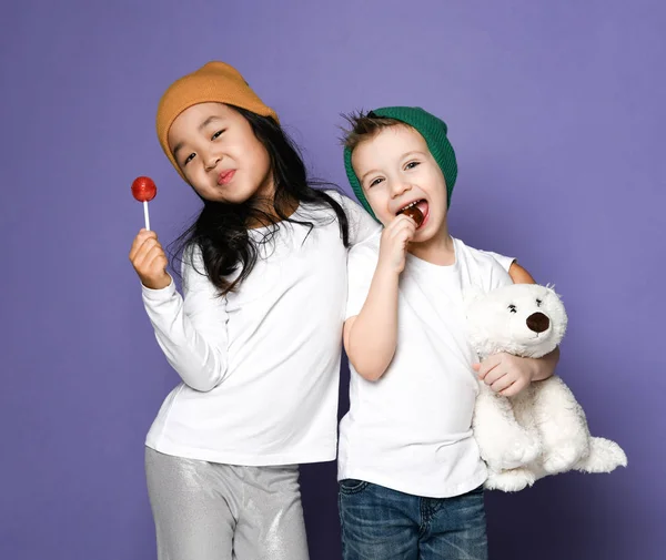 Два друга дети девочка и мальчик в белой футболке и красочные шляпы держат игрушку белого медведя и конфеты леденец счастливый улыбаясь на синей мяте — стоковое фото