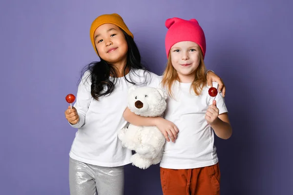 Beyaz t-shirt ve renkli pantolon ve şapka iki arkadaş çocuk kız kutup ayısı oyuncak ve lolipop şeker mutlu gülümseyerek tutun — Stok fotoğraf
