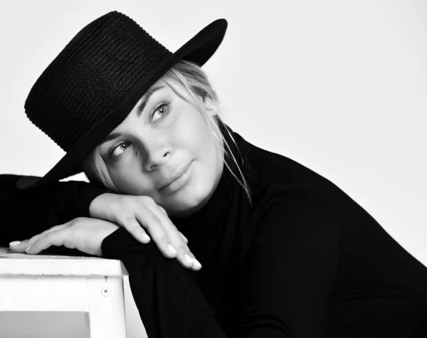 Black and white portrait of romantic woman in black hat with brim and turtleneck lying her head on her arms on white