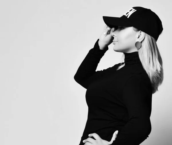 Black and white portrait of blonde woman in turtleneck holding the visor of her cap pulling it down over her eyes