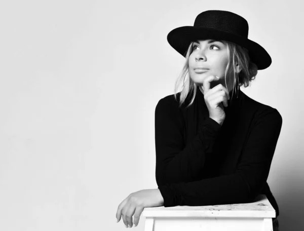 Black and white portrait of thoughtful woman in black hat with brim and turtleneck leaning her hands on the stepladder