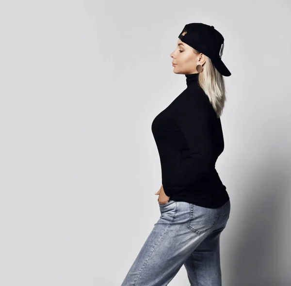 Snygg blondin kurviga kvinna i blå jeans, svart polotröja och mössa bärs bakåt poserar — Stockfoto