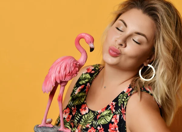 Portret van de blonde vrouw in sundress met tropische print Holding roze flamingo beeldje het verzenden van een kus op geel — Stockfoto