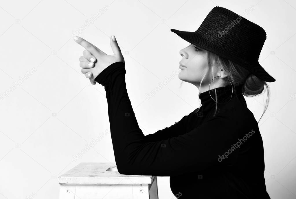 Black and white portrait in profile of blonde woman in black hat with brim doing a handgun gesture pointing at something